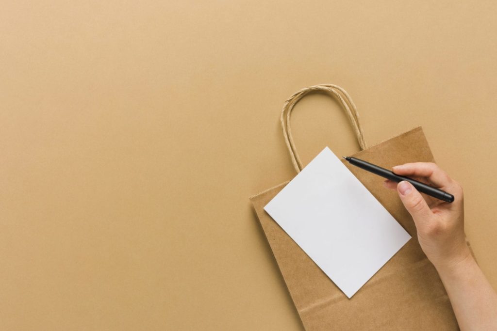 A white piece of paper is resting against a brown paper bag. There is a hand holding a pen above the paper. 